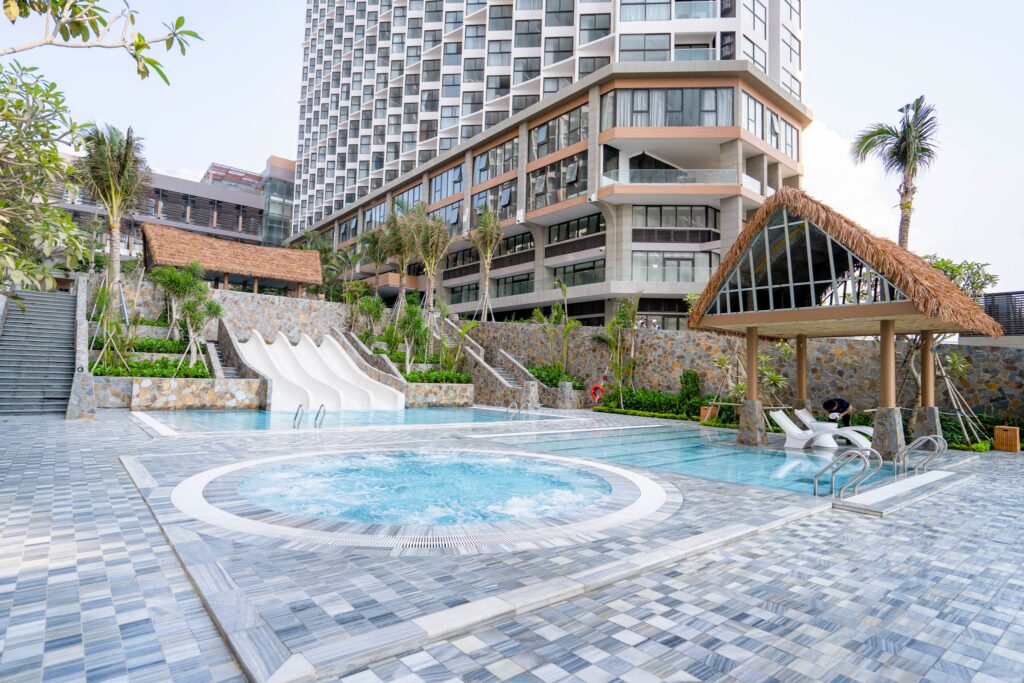 Mái Lá Nhân Tạo Bền Đẹp Cho Công Trình APEC Mũi Né 2025 2 Khu vực hồ massage và trượt nước trong resort với chòi nghỉ lợp mái lá nhân tạo cạnh tòa nhà cao tầng hiện đại
