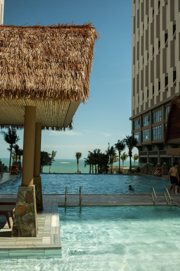 Mái Lá Nhân Tạo Bền Đẹp Cho Công Trình APEC Mũi Né 2025 3 Mái chòi hồ bơi resort lợp mái lá nhân tạo hướng biển với khung cột chắc chắn, bên cạnh tòa nhà cao tầng.