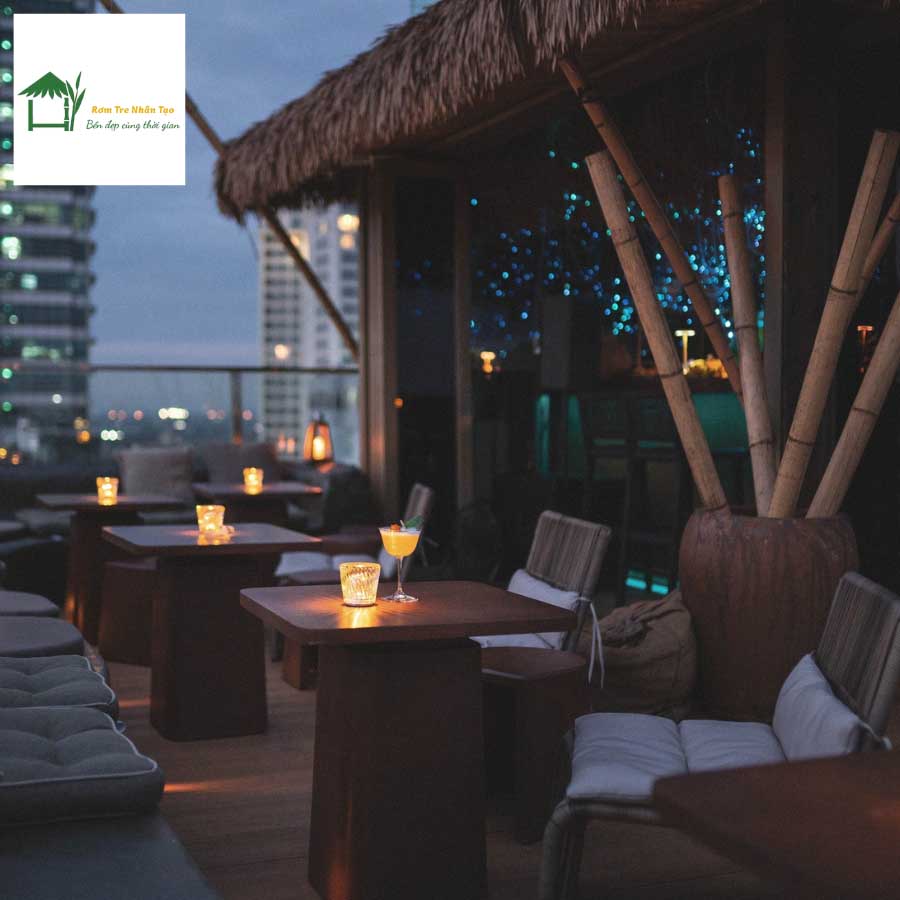 Góc cafe ngoài trời ban đêm, bàn ghế gỗ, đèn vàng, mái rơm nhân tạo, view cao tầng.